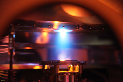 Photograph of the laser ablation process in the PLD chamber.The invisible, ultraviolet beam of an excimer laser hits a SrTiO3 target so that it shines as a blue disk (middle, top), creating a plasma plume (blue) which extends towards the substrate, a crystal of SrTiO3 (white rectangle in the middle, bottom).A second invisible infrared laser beam heats the substrate to 1200°C, so that it glows white.