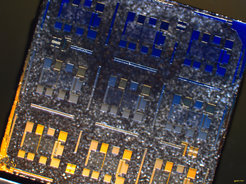 Optical micrograph of a 5x5mm LaAlO3-SrTiO3 sample containing nine devices which consist of FETs combined with Hall bars. Each device comprises a conducting channel of crystalline LaAlO3. The channel is well visible as a translucent area offset from the rest of the sample surface, which is covered with amorphous LaAlO3. The source and drain contacts and the voltage probes of the devices are provided by titanium films. The top-gates consist of PLD-grown gold. The diffuse, grainy background is due to the unpolished bottom face of the SrTiO3 substrate. Sample fabrication and micrograph: Lukas Kuerten.