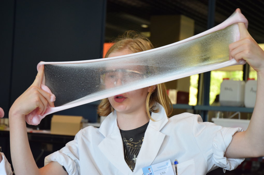 A pupil in a white lab coat stretches a translucent, glossy slime between their hands, showcasing its elasticity in a laboratory setting.