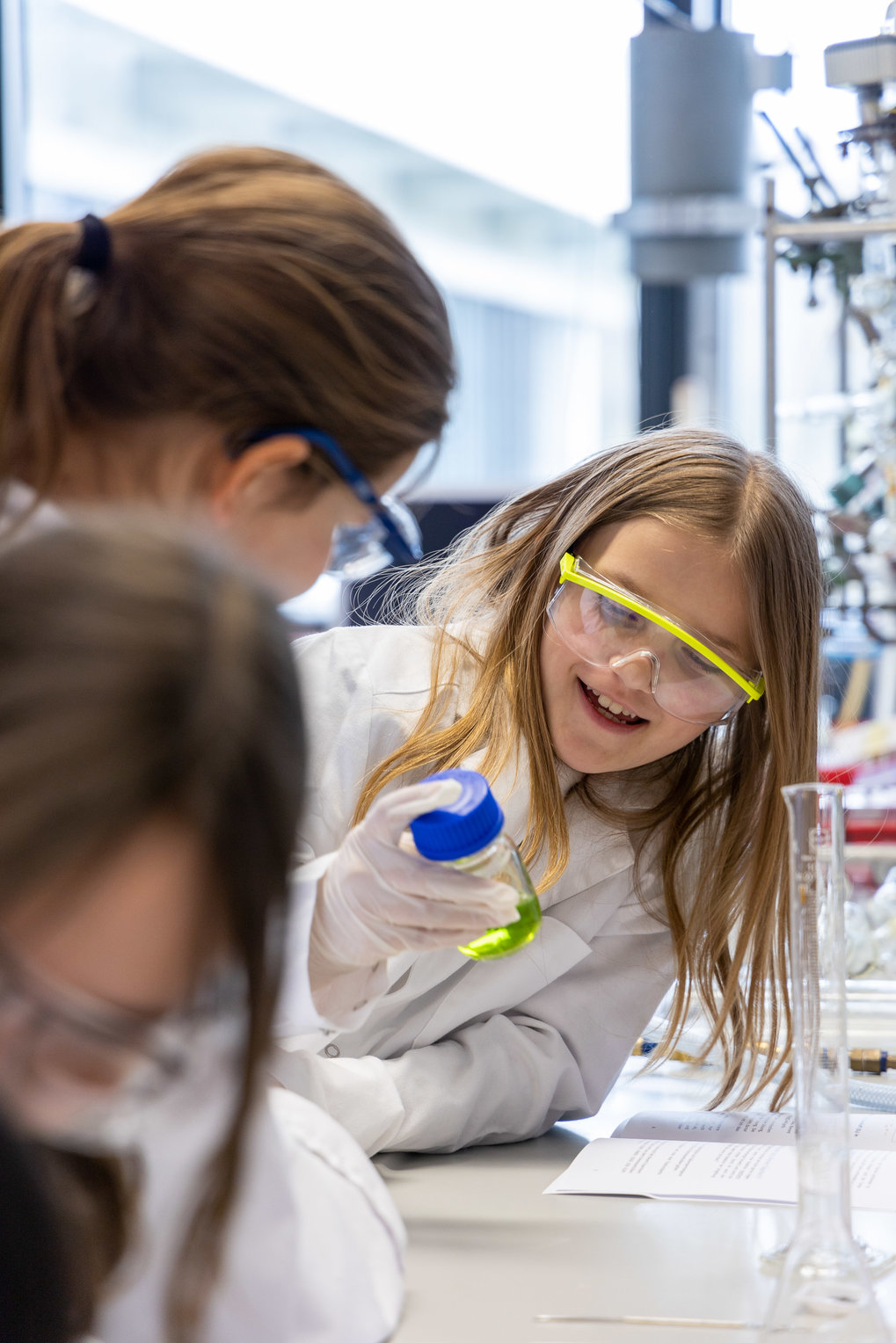 Pupils in white lab coats and lab goggles in the lab. One is holding a container with a yellow-green liquid