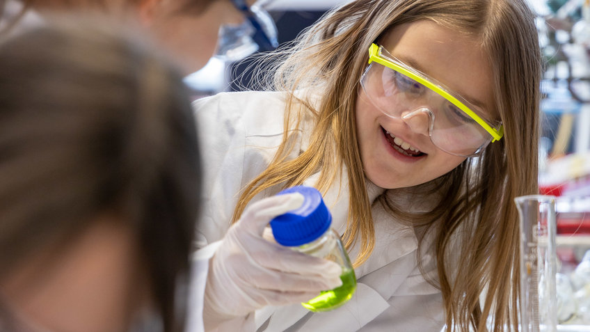 Girls’ Day Schülerinnen in weißen Laborkitteln und Laborbrillen im Labor. Einer hält einen Behälter mit einer gelb-grünen Flüssigkeit