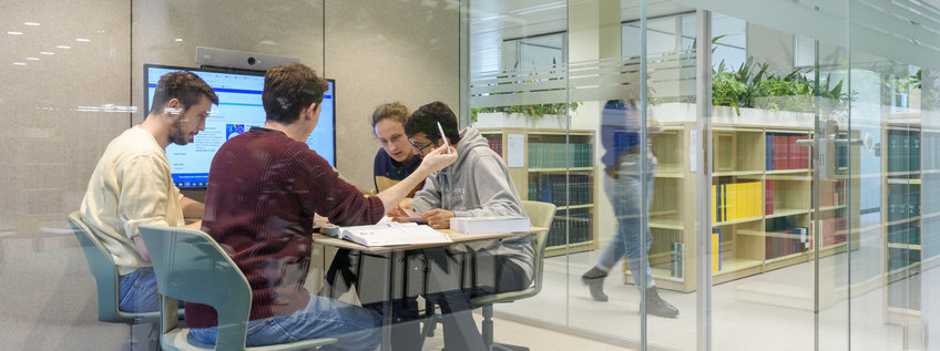Postdocs Vier Personen sitzen in einem modernen Raum mit Glaswänden, umgeben von Bücherregalen, und arbeiten zusammen. Hinter ihnen sind Bücherregale und eine vorbeigehende Person zu sehen. Ein Bildschirm im Raum zeigt zusätzliche Daten.