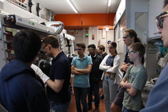 A group of people stands in a laboratory environment, focused on a large, complex piece of scientific equipment. The room is well-lit and features white and orange accents, suggesting a modern research setting. Various lab apparatus and safety gear are visible, indicating an active scientific workspace.