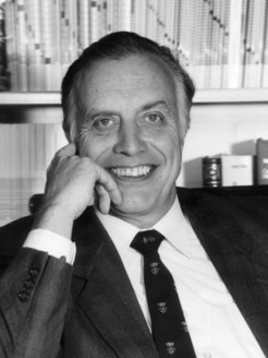 Prof Queisser dressed in a suit and patterned tie is seated in his office, surrounded by a bookshelf containing various books, indicating a professional setting.