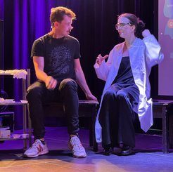 A stage scene with two individuals; one wears a black t-shirt and sneakers, the other a white lab coat, seated against a backdrop of purple curtains.