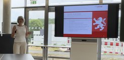 Indoor presentation showing a speaker Ms. Tanja Desch near a screen with "ProLOEWE" initiatives. The slide includes bullet points and a red lion symbol, with a view of an outdoor area through large windows.