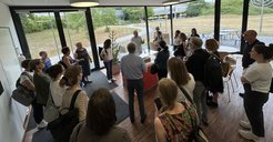 Participants of the SciComm standing in a well-lit room gathered closely around a central table with a Precision Building Modell. Dr Ast is giving a talk about the building history and its use.